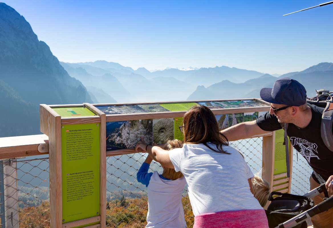 Eine Familie lernt interessante Dinger über die Region an einer Lernstation vor beeindruckendem Alpenpanorama bei bestem Wetter.